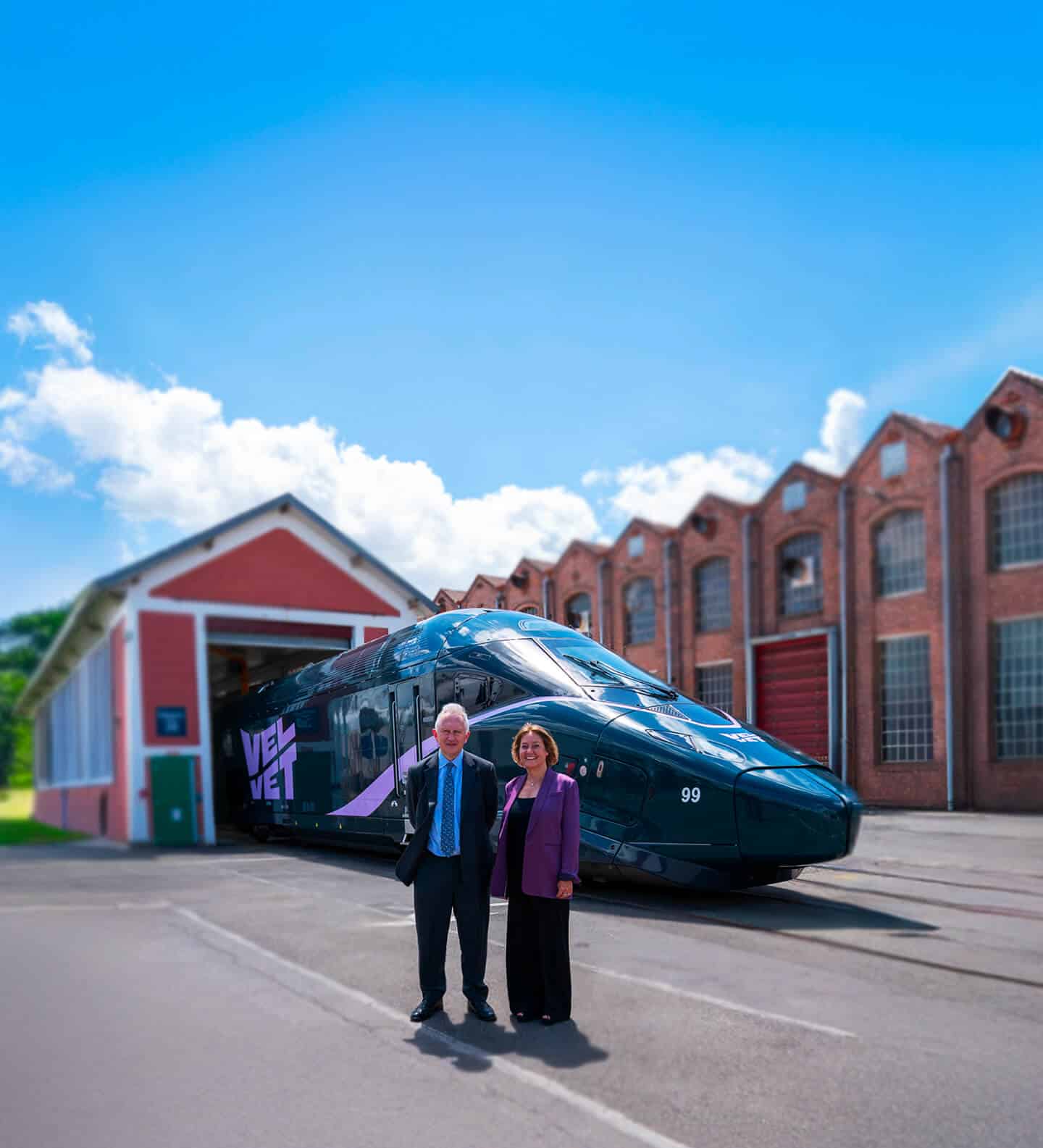 Vue d'ensemble du nouveau train "VELVET", flanqué de deux dirigeants devant un hangar et des bâtiments industriels en briques rouges sous un ciel bleu éclatant.